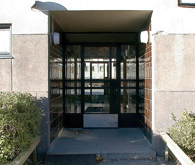Stockholm, Bredäng, Coldinuorden 3, Vita Liljans väg 30-40. En av byggnadens genomgående entréer, fotograferat från öster. Notera dörrpartiets smäckra ramverk av svart stål. 
