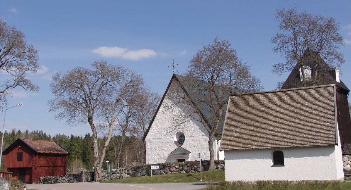 En slingrande väg passerar kyrkan med omgivande  kyrkogårdsmur, tiondebod och bårhus, samt prästgården från 1880-talet på andra sidan.
