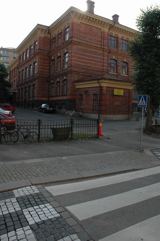Nya Elementarläroverket för flickor, baksidans fasad mot Gustavsgatan, kortsidan mot Vasagatan. Skolgården omges av gjutjärnsstaket och planterade lövträd.