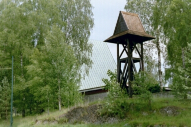 Här ses Sjöbo kyrka från Fristadvägen, höljd i grönska, med sin äldre klockstapel i förgrunden.