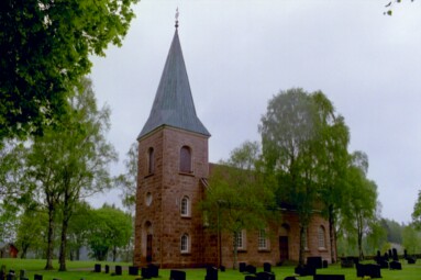 Björkar flankerar gångarna runt Vänga kyrka och innanför kyrkogårdsmurarna står i huvudsak lönnar. Till vänster i bild syns en minnessten över den gamla kyrkan och vapenhuset.