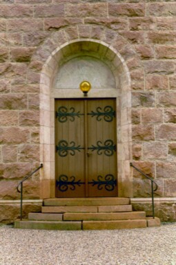 Båda portarna till Vänga kyrka har likadan utformning. Här ses den västra, där inskriptionen anger byggnadsåret, 1905. De stora smidesbeslagen är typiska för nationalromantikens vurm för den medeltida byggnadstekniken, som rådde vid uppförandet.