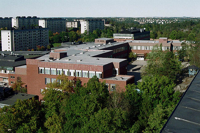 Skolans huvudbyggnad med den mot öster öppna gården, sedd från taket på ett närbeläget skivhus. Till höger i bilden anas det stora utsparade naturmarkspartiet som upptar mittdelen av skolgården. I bakgrunden skivhusen i kv. Gula Knapparna och Amarantern. 
SAK00042_Stockholm, Bredäng, Concordia 4 och 5, Bredängstorget 13-25,27,29,Concordiavägen 7-25_från syd
